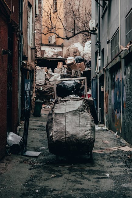 A large, rusty red metal dumpster with vertical ridges on its sides is positioned against a dark grey wall on the left side of the image. The dumpster shows signs of weathering and paint chipping, wit