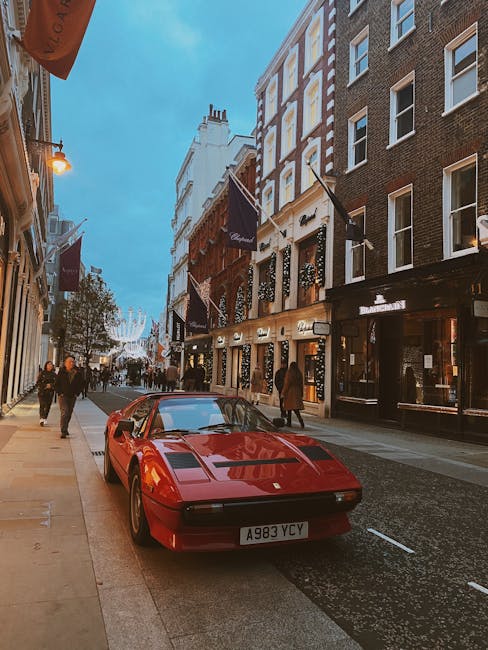 A red sports car with a sleek, low-profile design and smooth, shiny finish is parked on the side of a bustling city street, adjacent to a beige-colored curb. The car features a distinctive aerodynamic
