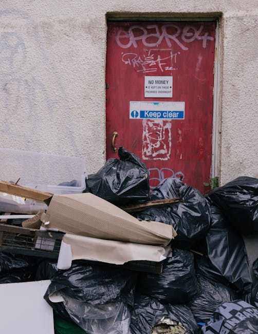 In the foreground, a light blue refuse collection vehicle is parked on a city street, situated to the right of a gated entrance flanked by stone pillars and greenery. The vehicle appears to be in an i